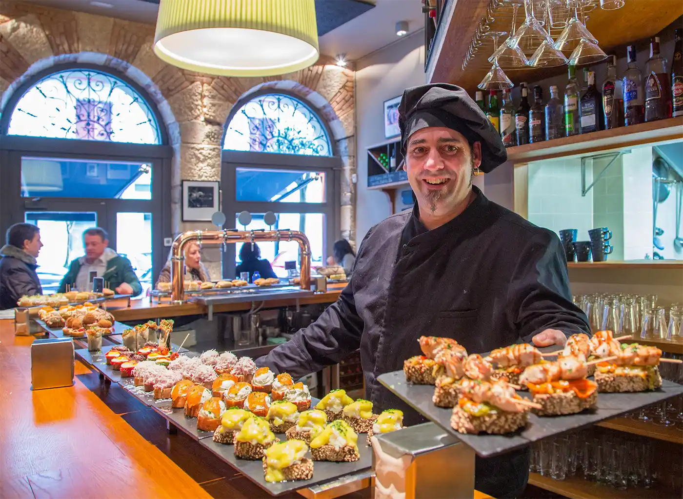 Bilbao-Pintxos-Bar-Kellner Kellner mit schwarzer Mütze zeigt Pintxos auf zwei Platten in einem Restaurant