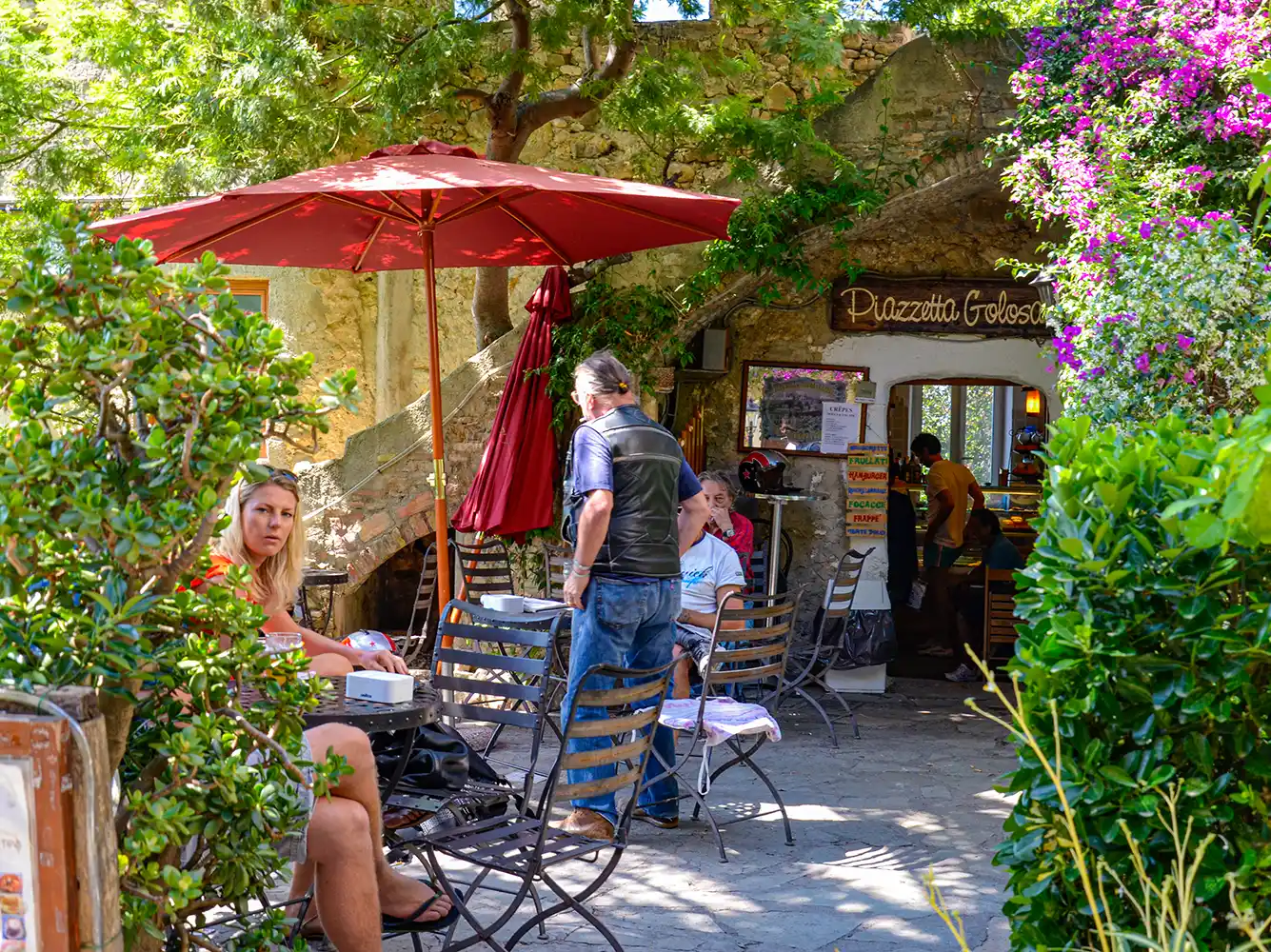 Menschen im Sommer an Tischen unter einem roten Sonnenschirm vor einem Restaurant
