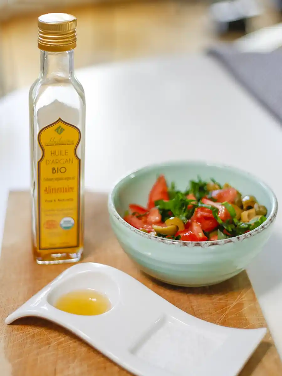 Eine Flasche Arganöl mit einem kleinen Schälchen und einer Schüssel Salat mit Tomaten und Oliven auf einem Schneidebrett.