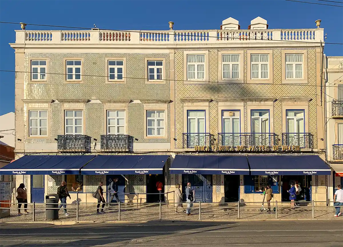 Ein sonnenbeschienenes, gekacheltes Gebäude mit blauen Markisen und Menschen, die auf dem Gehweg davor spazieren gehen.
