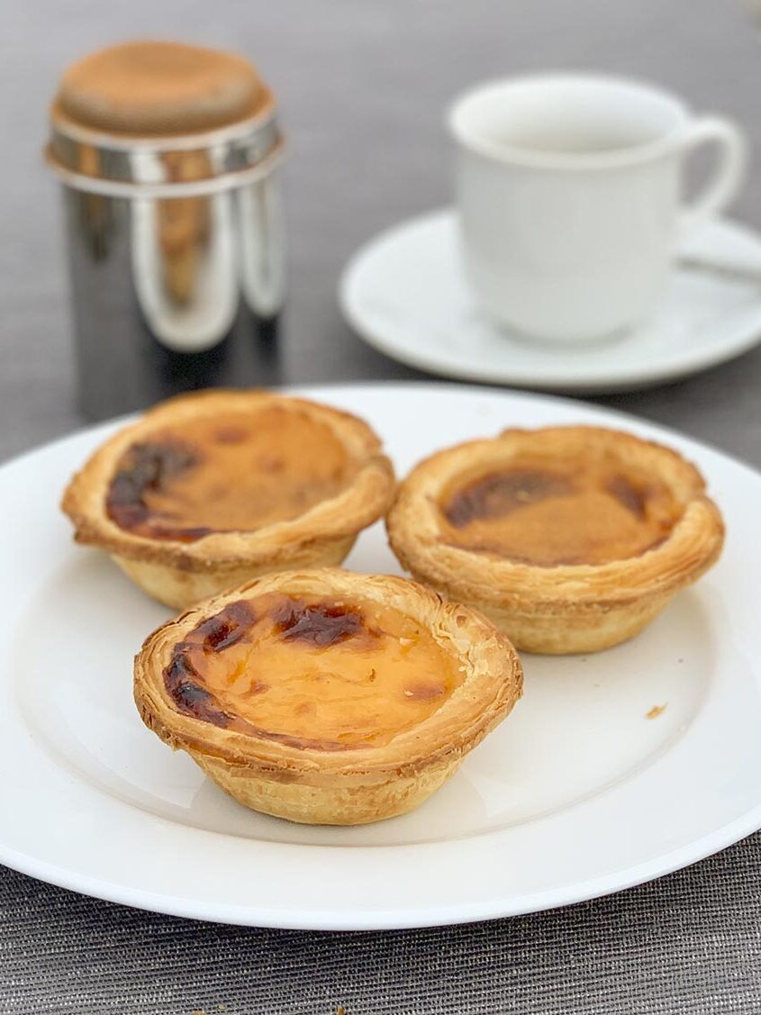 3 pasteis de nata, im Hintergrund ein Espresso und Zimtstreuer