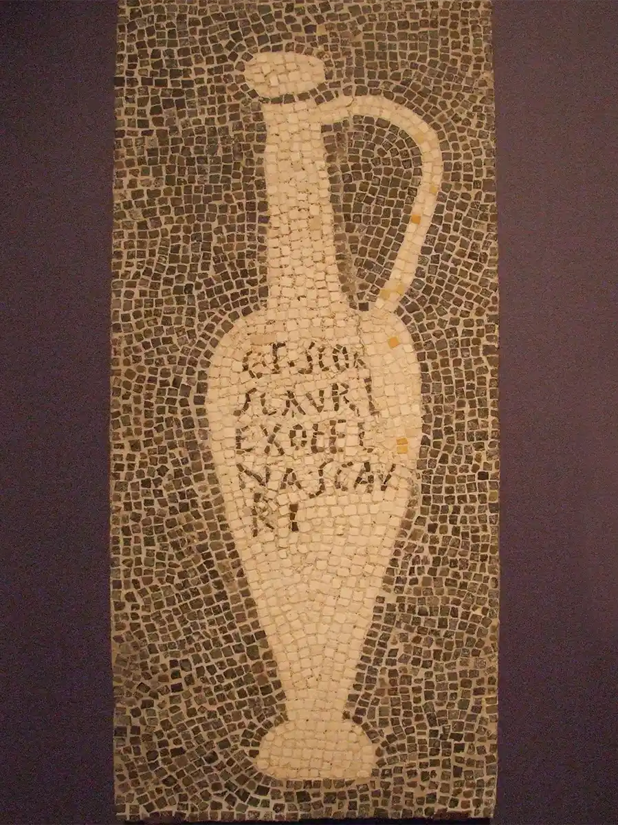 Antikes Mosaik eines hohen Kruges mit Henkel, mit lateinischem Text auf dem Korpus, vor dunklem Hintergrund.