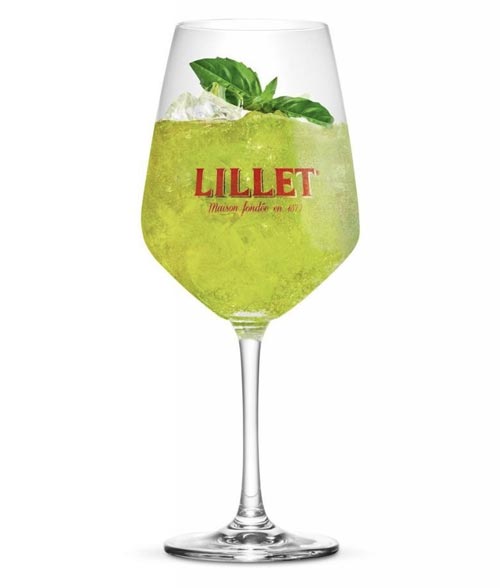 Ein Weinglas mit grünem Cocktail, Eis, Basilikumblättern und Lillet-Branding auf dem Glas.
