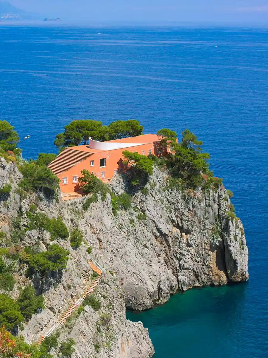 Ein Haus aus Terrakotta steht auf einer felsigen Klippe mit Blick auf das blaue Meer, umgeben von Bäumen und Grünflächen.