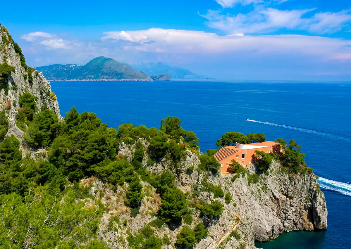 Eine felsige Küste mit üppigem Grün, ein Haus auf einer Klippe und blaues Meer unter hellem, klarem Himmel.