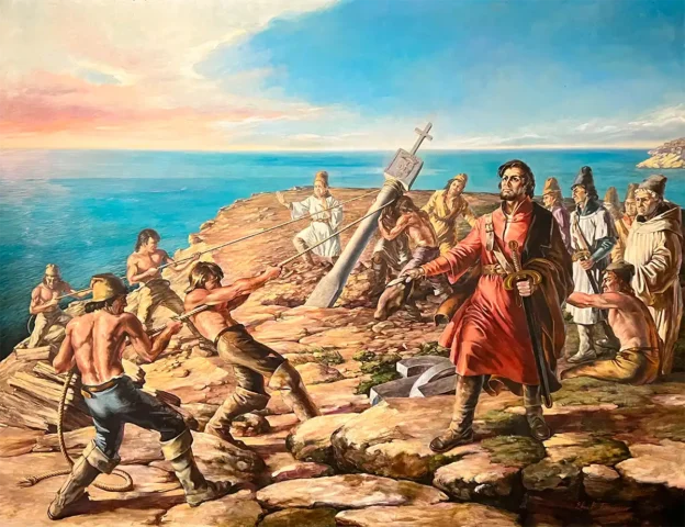 Männer in historischer Kleidung errichten ein steinernes Kreuz an einer felsigen Küste unter blauem Himmel mit dem Meer im Hintergrund.