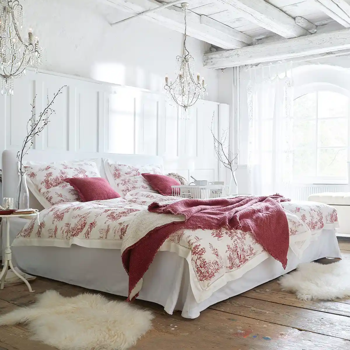 Helles Schlafzimmer mit weißen Wänden, Holzböden, Kronleuchtern und einem Bett mit roter und weißer Toile de Jouy Bettwäsche und Kissen.