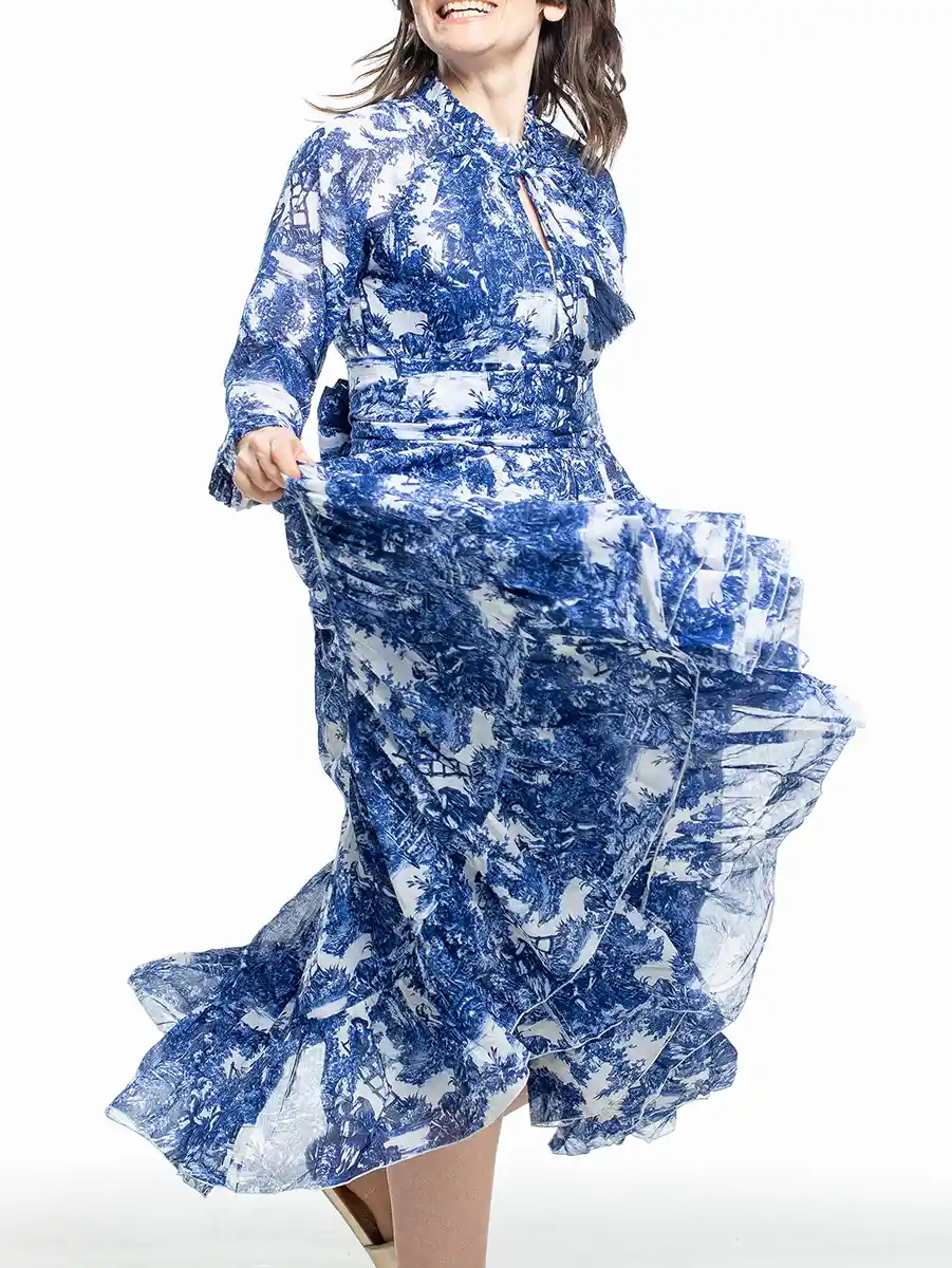 Frau in einem fließenden langärmeligen Kleid aus blauem und weißem Toile-de-Jouy, das sich vor einem schlichten weißen Hintergrund dreht.