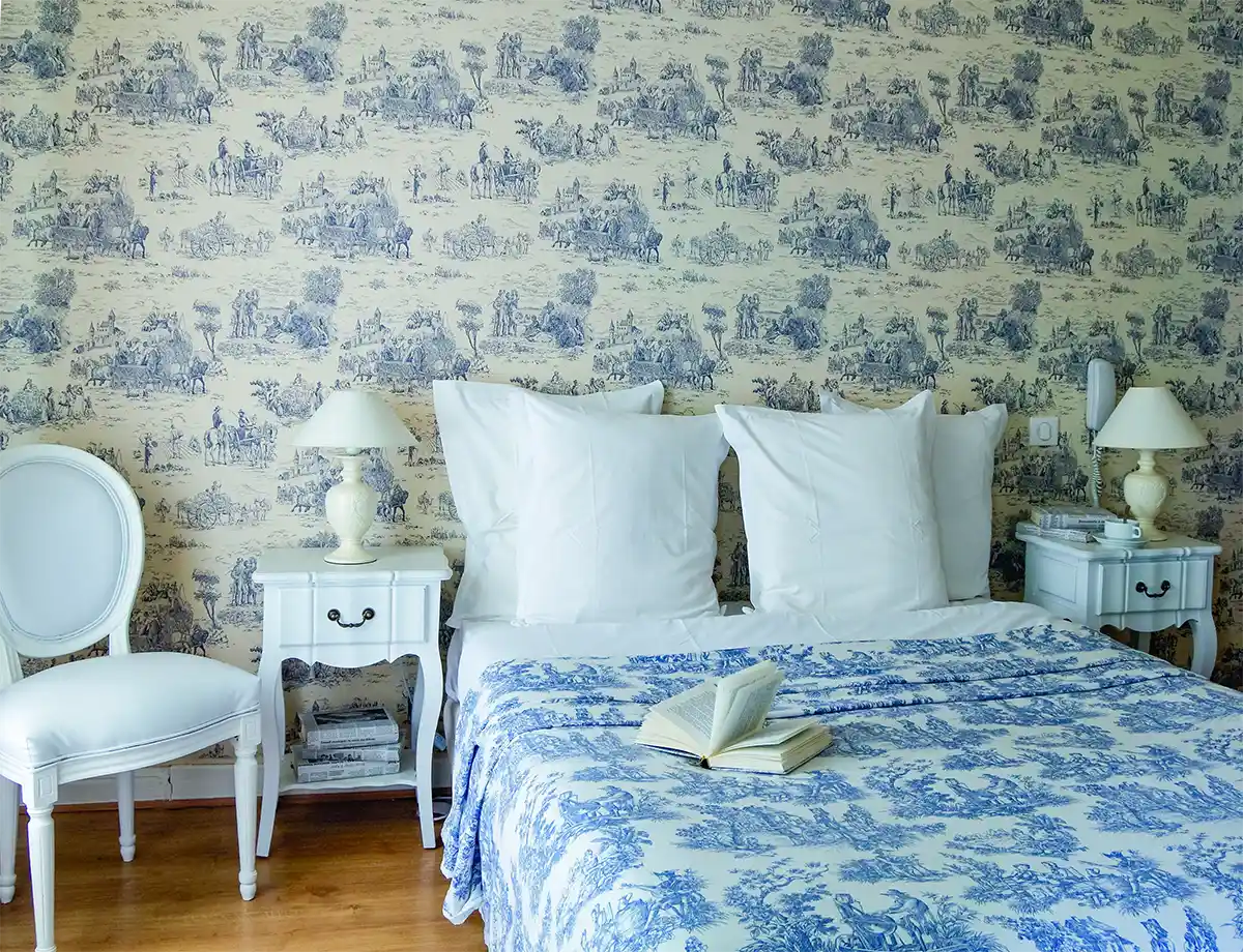 Elegantes Schlafzimmer mit blauem und weißem Dekor im Toile de Jouy-Stil gemusterten Tapeten, und Bettdecke, Nachttischen, Lampen und einem offenen Buch.