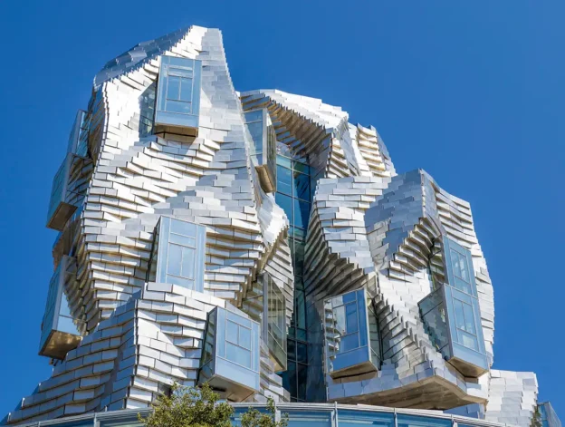 Das Luma in Arles: Modernes Gebäude mit glänzenden, unregelmäßigen Metallpaneelen und hervorstehenden Fenstern vor einem klaren blauen Himmel.