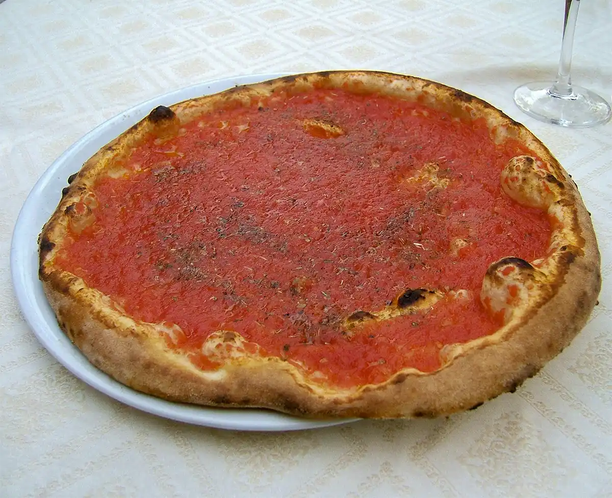 Original-Pizza-Marinara Eine einfache Pizza mit Tomatensoße auf einem weißen Teller, der auf einem gemusterten Tischtuch liegt.