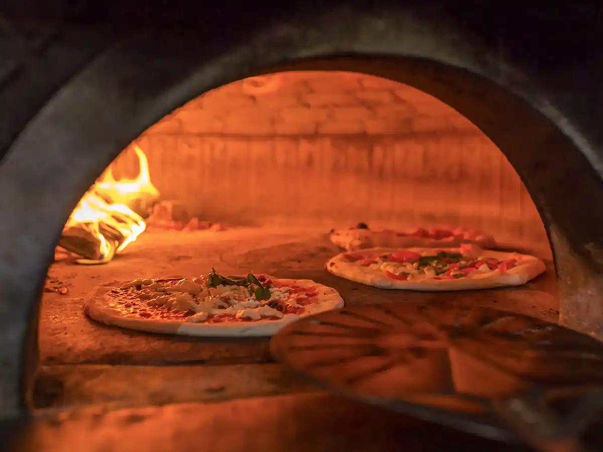 Pizza-Pizzaofen-Neapel Holzofen mit Flammen, der drei mit Käse, Tomaten und Basilikum belegte Pizzen auf einer Steinfläche backt.