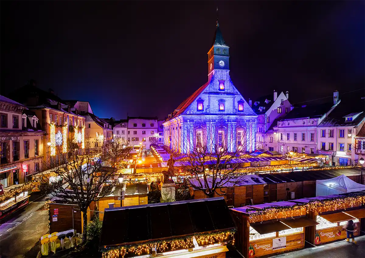 Ein festlicher Weihnachtsmarkt mit beleuchteten Ständen und einer nachts blau beleuchteten Kirche.