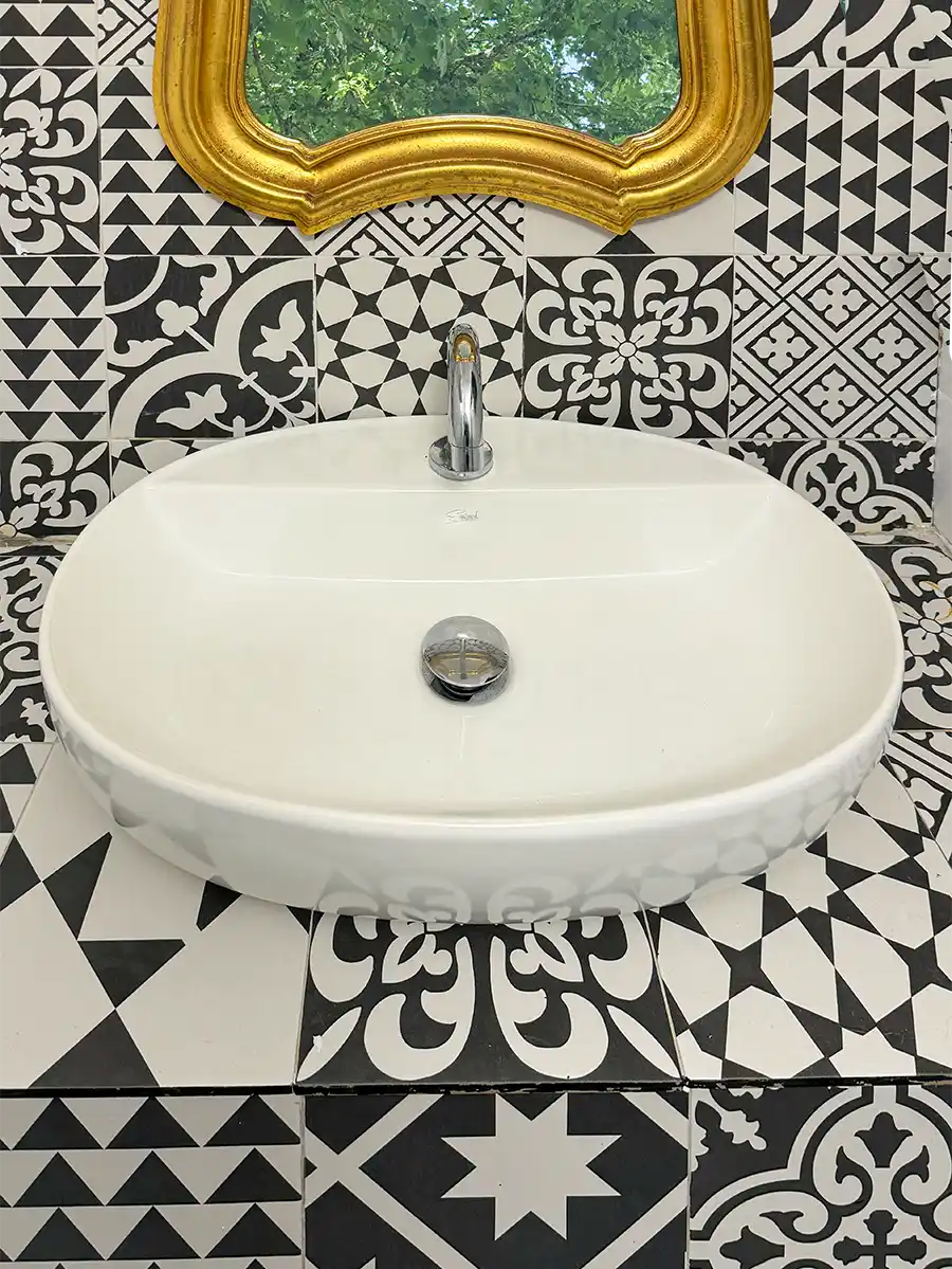 Badezimmer-mediterran-Azulejo-Optik Weißes ovales Waschbecken mit verchromtem Wasserhahn vor schwarz-weiß gemusterten Kacheln und einem goldgerahmten Spiegel.