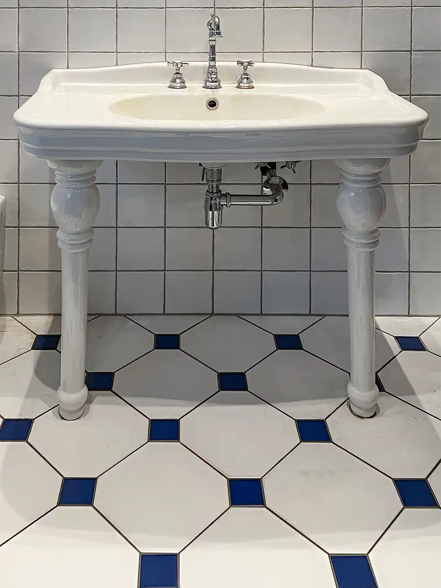 Badezimmer-mediterran-Fliesen Ein weißes Porzellanwaschbecken mit verchromten Armaturen und zwei Füßen, vor weiß gefliesten Wänden und Böden mit blauen Akzenten.