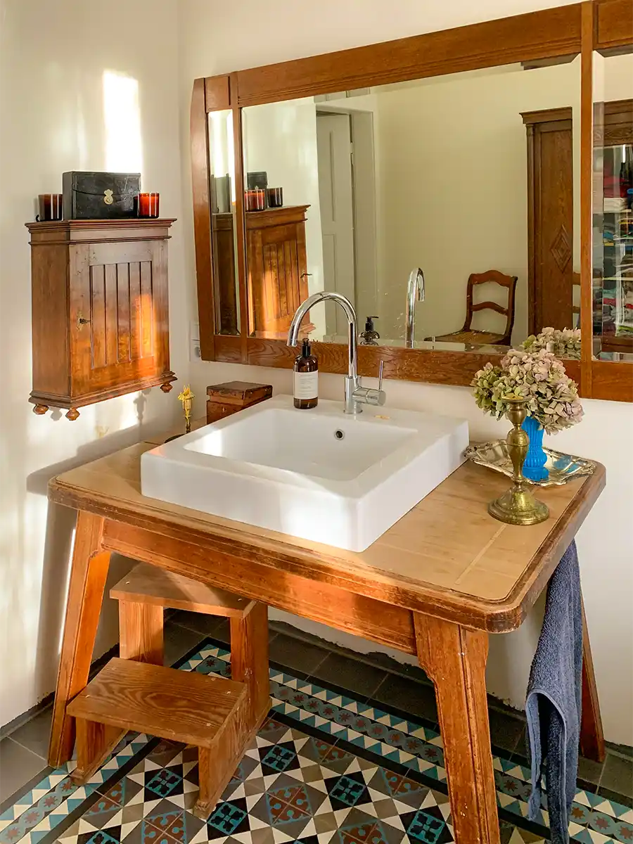 Badezimmer-mediterran-Holztisch Badezimmer im Vintage-Stil mit hölzernem Waschtisch, quadratischem Waschbecken, Blumen, Spiegel und dekorativen Bodenfliesen.