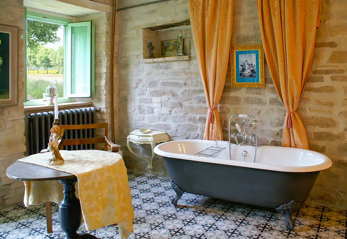 Badezimmer-mediterran-Italien-Bauernhof Vintage-Bad mit Krallenfußwanne, gelben Vorhängen, gemustertem Fliesenboden und offenem Fenster mit Grünpflanzen.