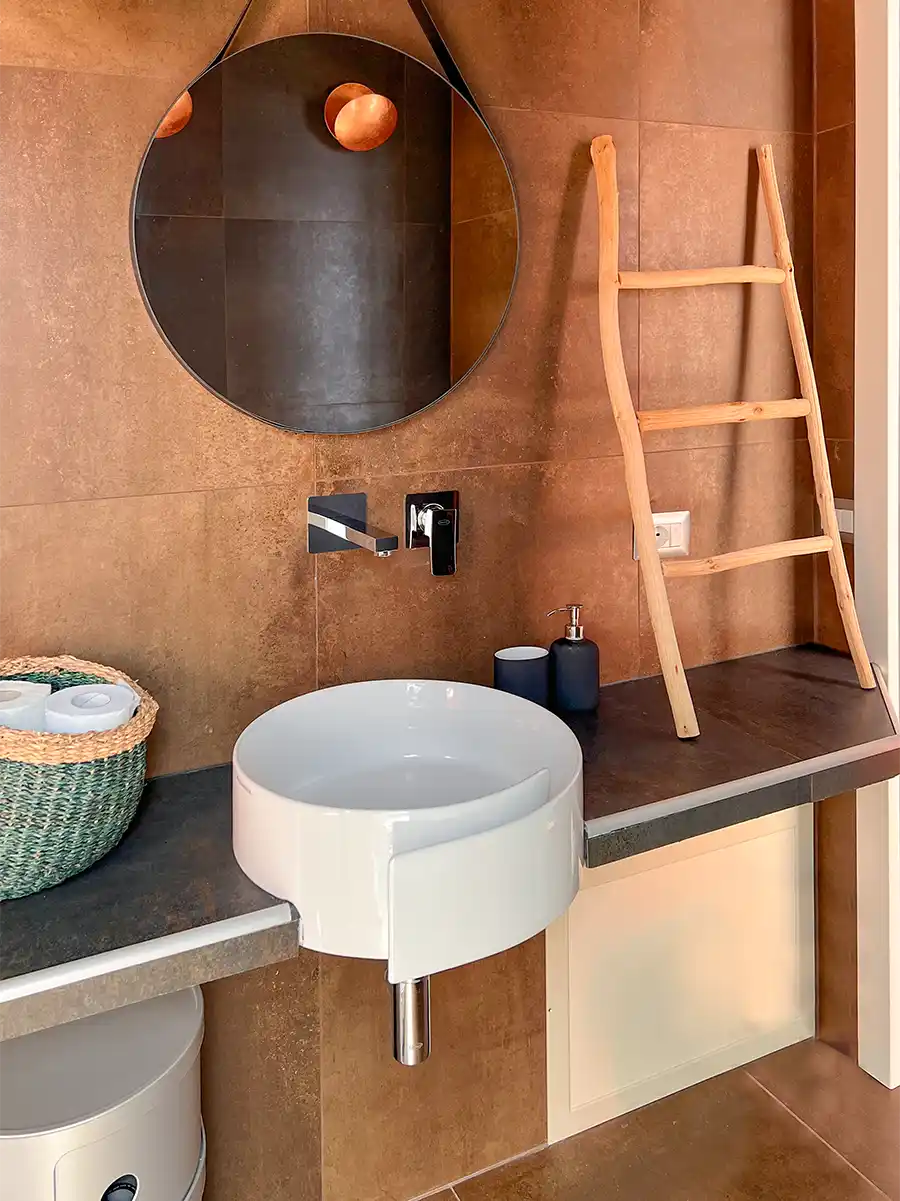 Badezimmer-mediterran-warme-Farben Modernes Badezimmer mit rundem Spiegel, Gefäßwaschbecken, Seifenspender und einer Holzleiter vor einer braunen Wand.