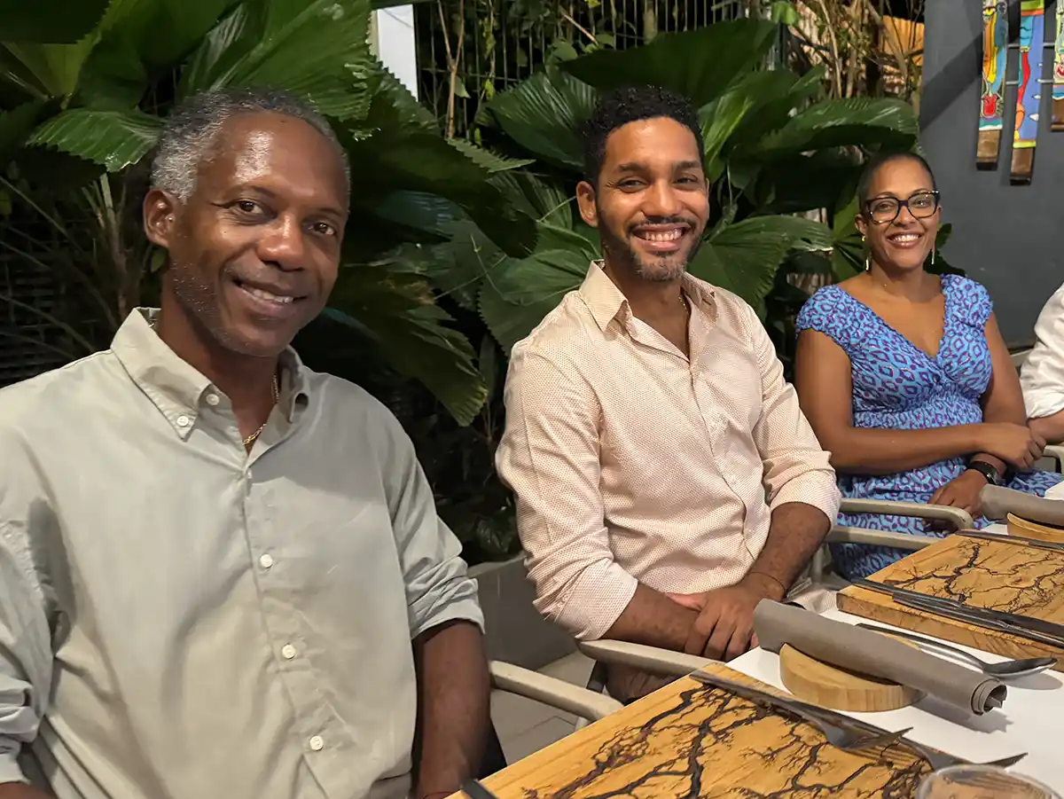 Martinique-Coderum Drei Personen sitzen an einem Tisch im Freien und lächeln in die Kamera.