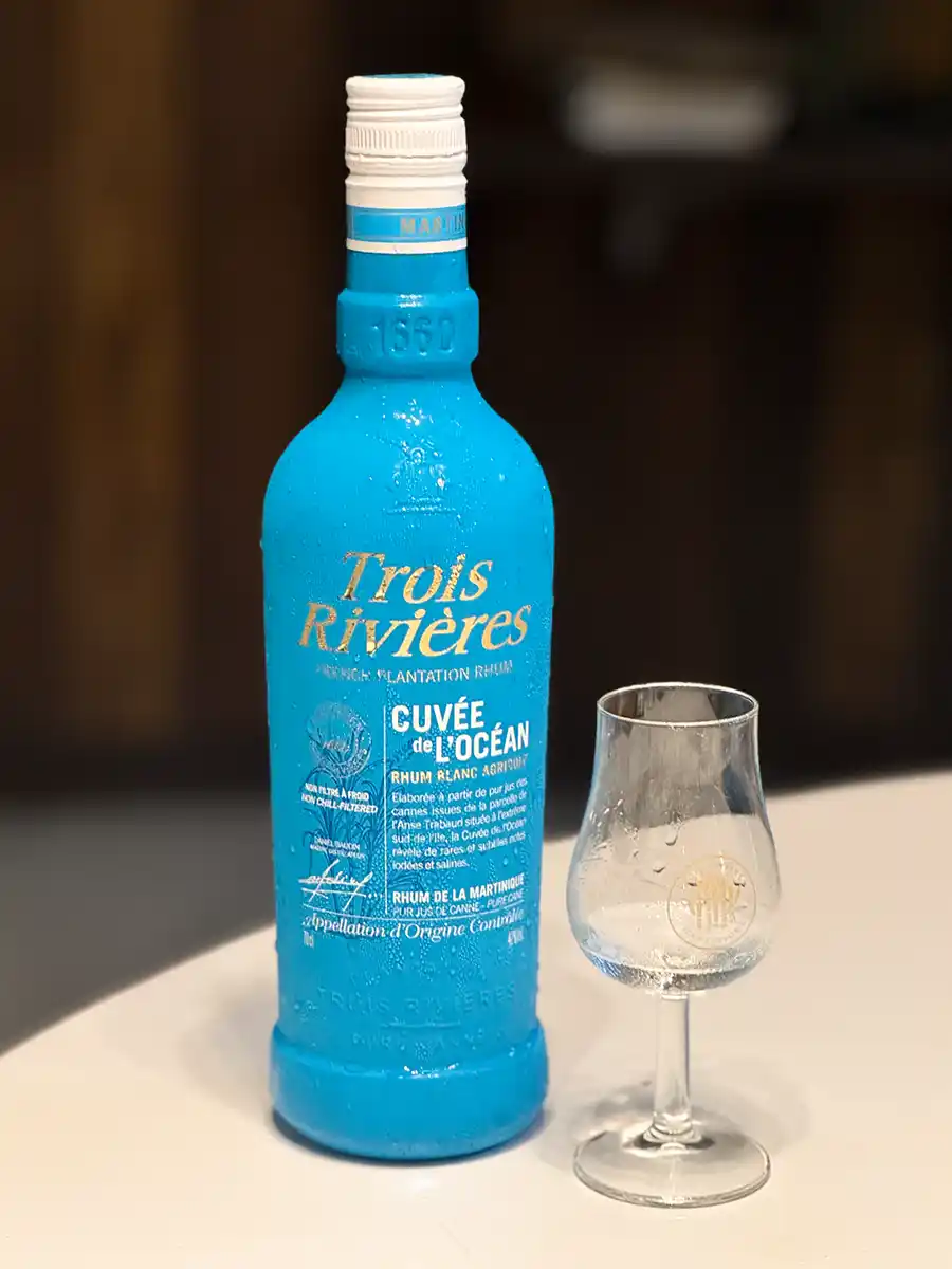 Martinique-Rum-Agricole-Trois-Rivieres Eine blaue Rumflasche von Trois Rivières steht neben einem leeren Glas auf einer weißen Fläche.