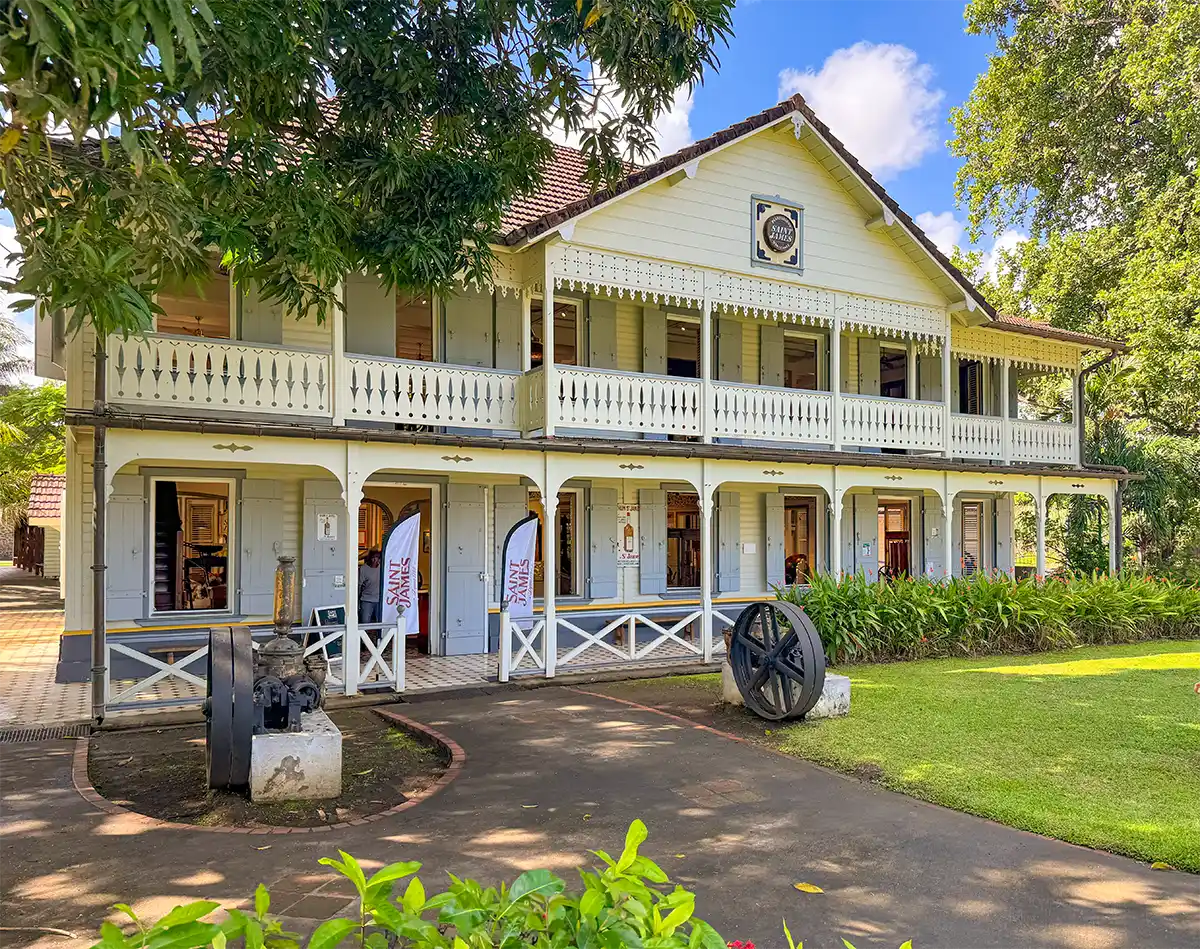 Martinique-Rum-Museum-Saint-James Zweistöckiges Gebäude im Kolonialstil mit einer Veranda, weißem Geländer und einer Rasenfläche unter blauem Himmel.