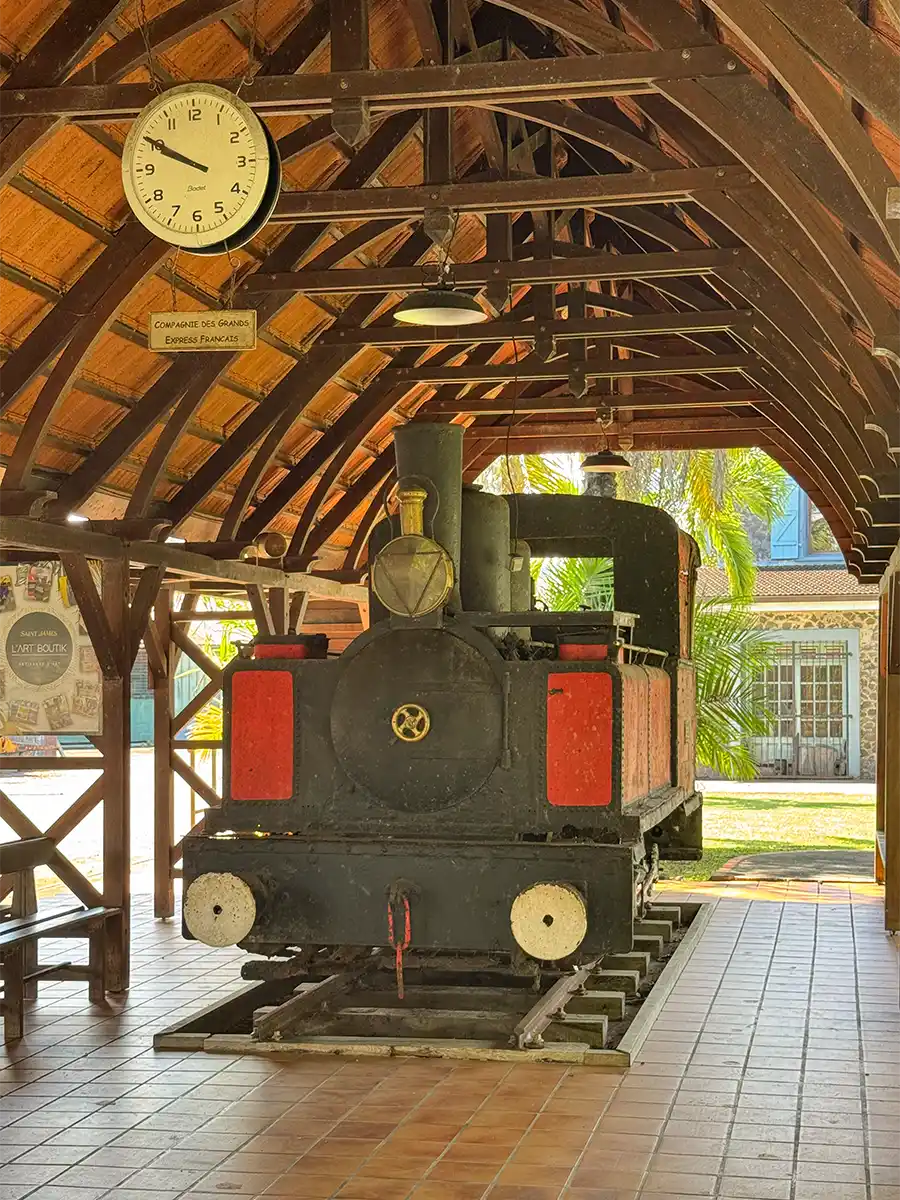 Martinique-Zuckerrohr-Eisenbahn Eine alte schwarze Lokomotive ist in einem Innenraum unter einem Holzdach ausgestellt, über dem eine Uhr hängt.