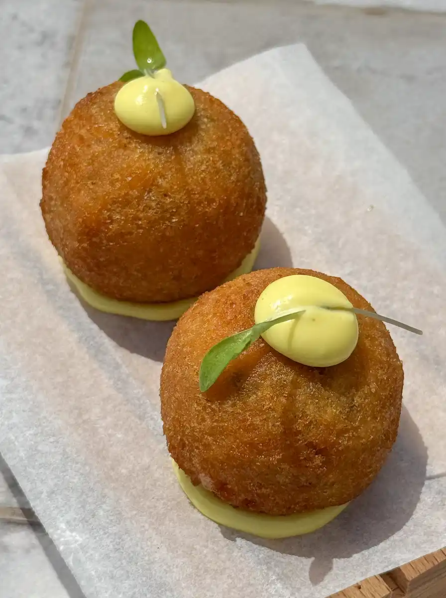Zwei runde, goldbraune Kroketten, belegt mit gelber Sauce und einem kleinen Blatt auf Pergamentpapier.