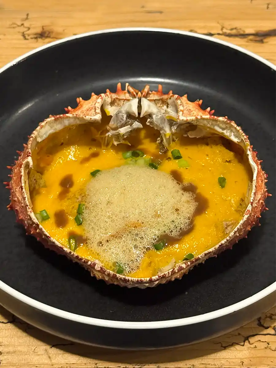 Rum-Food-Pairing Eine Krabbenschale gefüllt mit gelber Sahnesauce, Schaum und garniert mit gehackten grünen Kräutern auf einem schwarzen Teller.