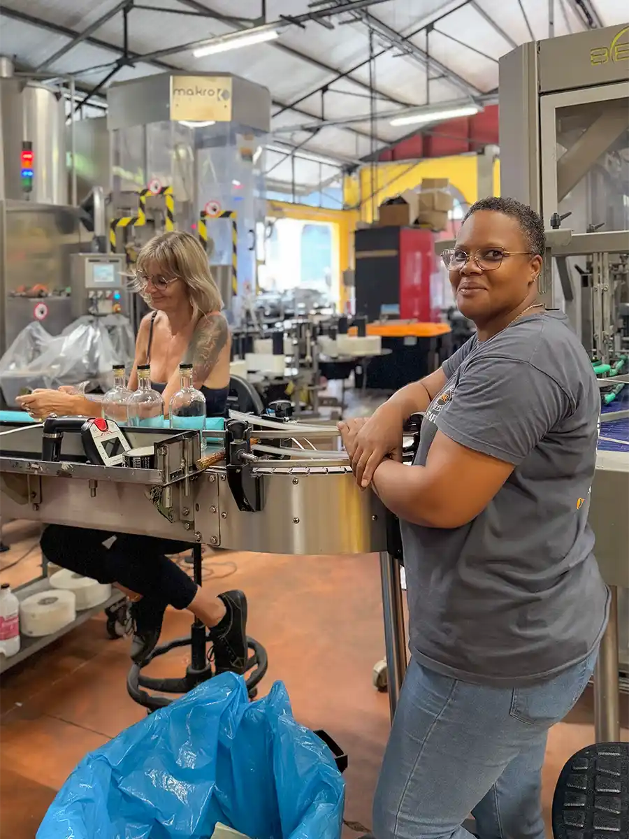 Rum-HSE-Qualitaetspruefung Zwei Frauen arbeiten in einer Fabrik; eine sitzt und etikettiert Flaschen, die andere steht und lächelt in die Kamera.