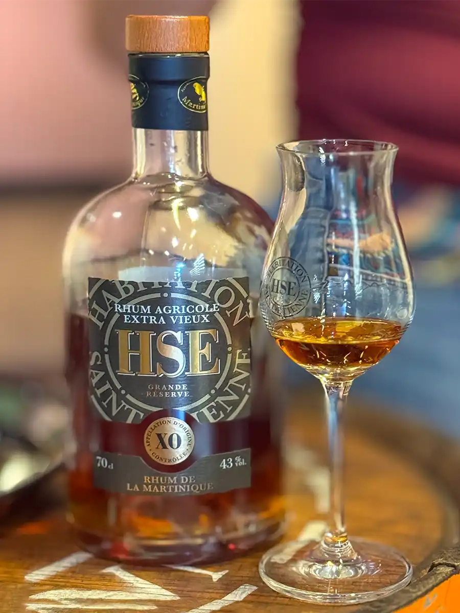 Rum-HSE-XO-Extra-Old Eine Flasche HSE-Rum und ein Glas mit bernsteinfarbener Flüssigkeit auf einem Holztablett.