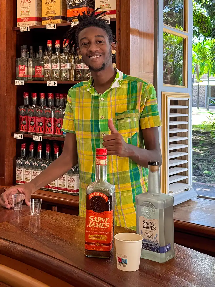 Rum-Museum-Saint-James-Joshua-Bar Ein lächelnder Mann in einem gelbkarierten Hemd zeigt hinter einer Bar mit Saint-James-Flaschen den Daumen nach oben.