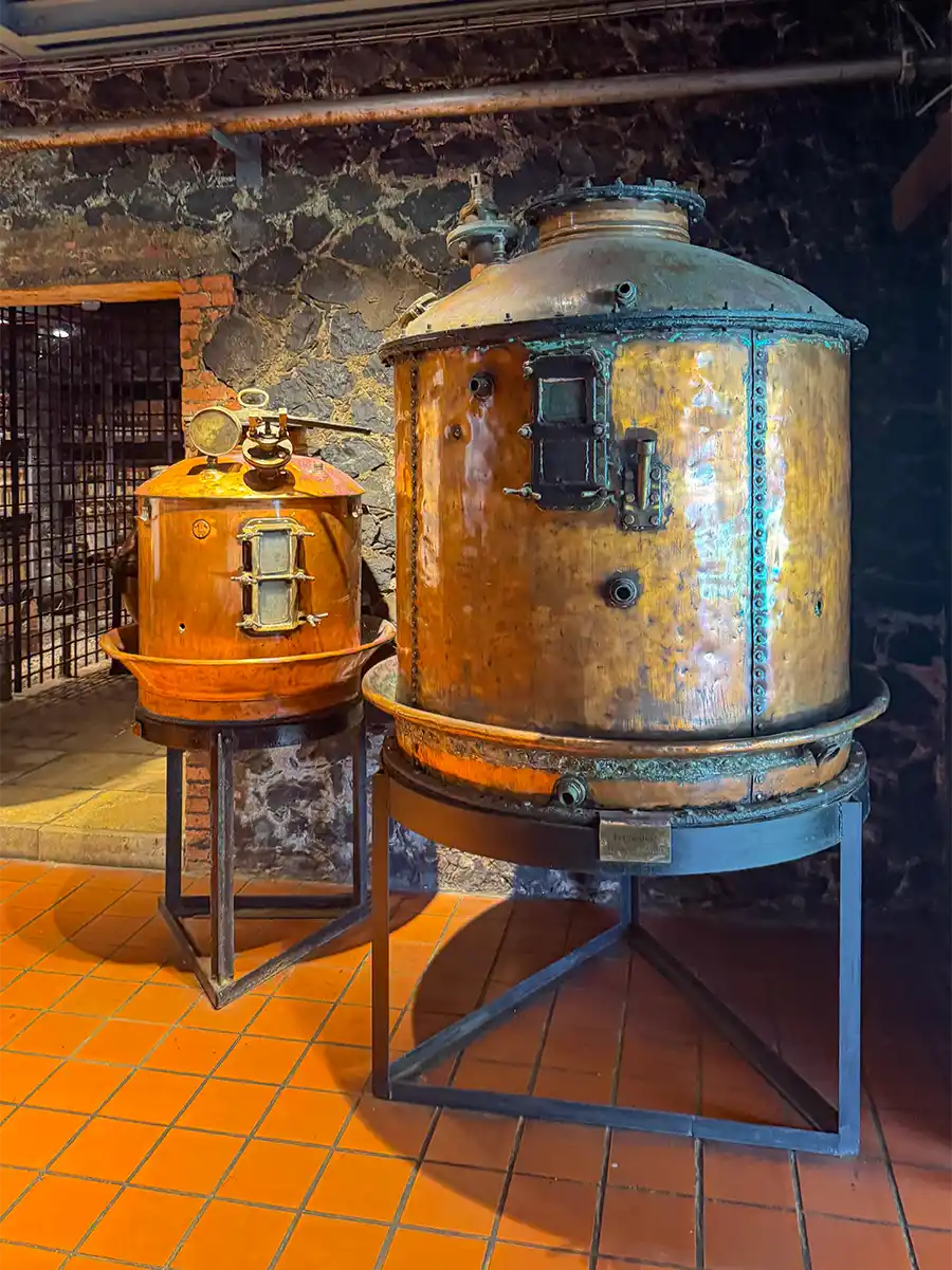 Rum-Museum-Saint-James-Kupferkessel Zwei große Destillationstanks aus Kupfer auf Metallständern in einem rustikalen Raum mit Steinwänden und Fliesenboden.