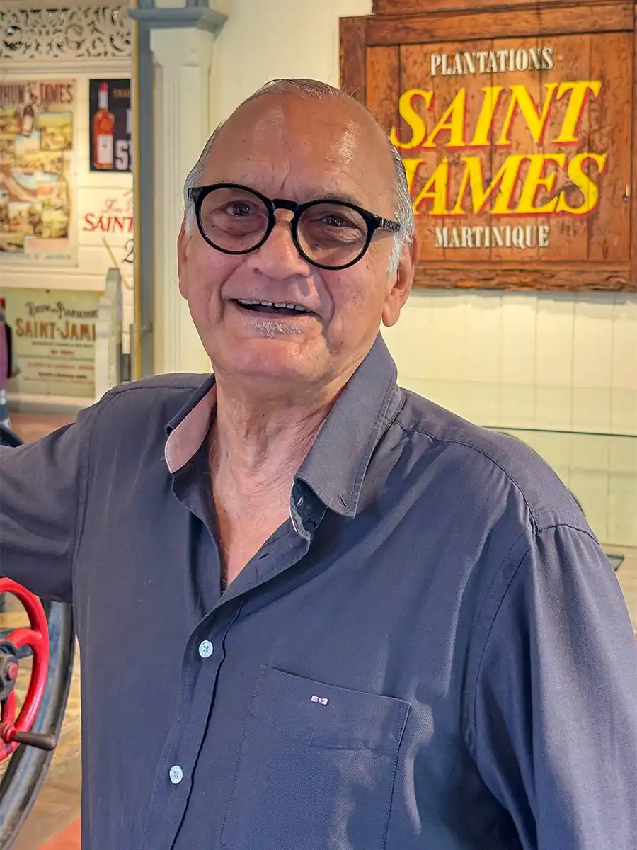 Rum-Museum-Saint-James-Michel-Fayad Ein lächelnder älterer Mann mit Brille und grauem Hemd steht in einem Haus neben einem Schild von Saint James Martinique.