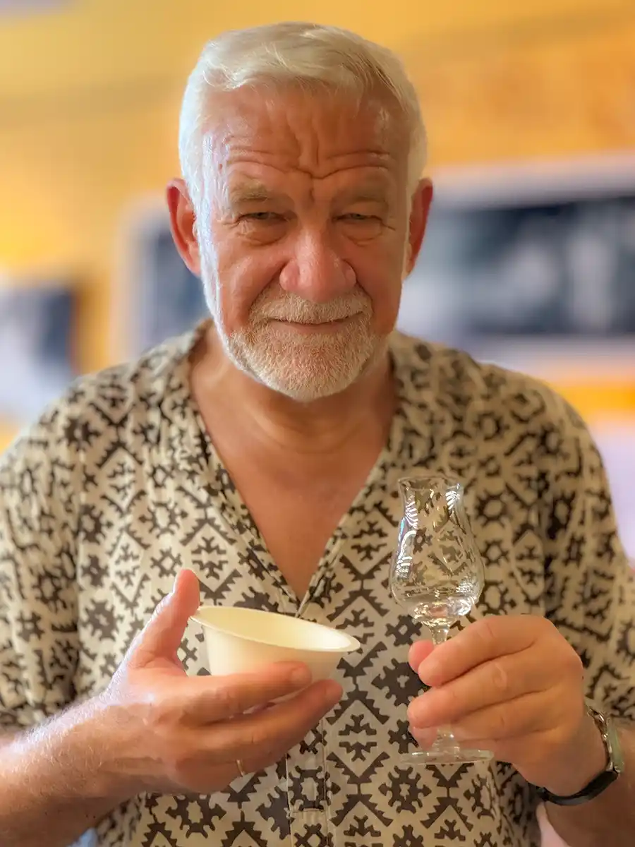 Rumverkostung-Siegbert-Mattheis Ein älterer Mann mit weißem Haar hält ein kleines Glas und eine Papierschale in der Hand und lächelt in einer warm beleuchteten Umgebung.