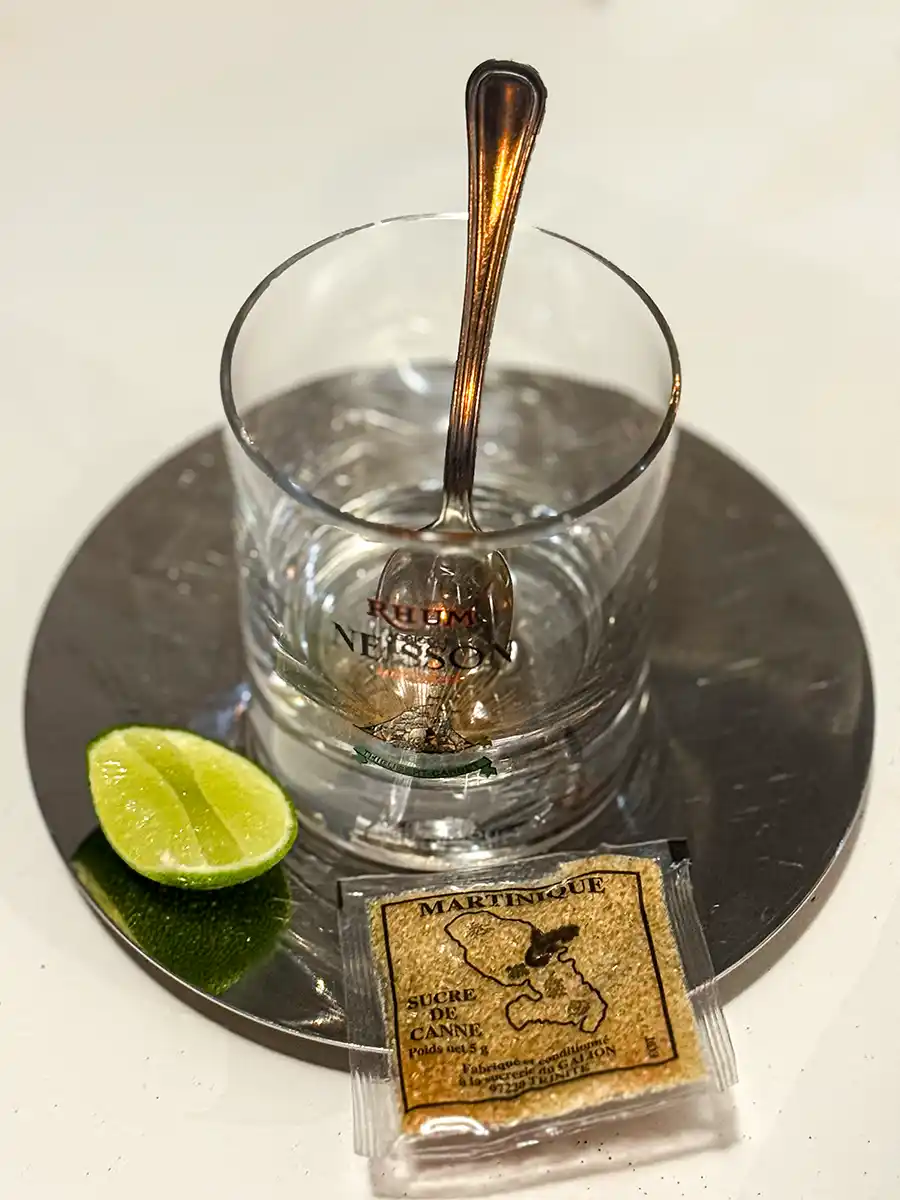 Ti-Punch-Martinique Ein leeres Glas mit einem Löffel, einer Limettenspalte und einem Zuckerpäckchen auf einem runden Metalltablett.