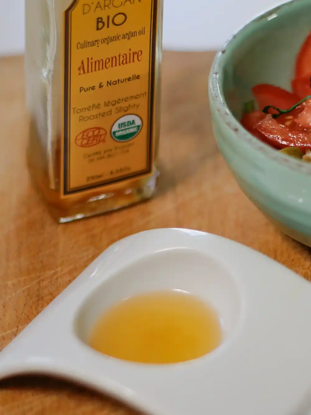 Eine kleine Schale mit Arganöl neben einer Flasche mit kulinarischem Arganöl und einer Schüssel mit Salat auf einer Holzfläche.