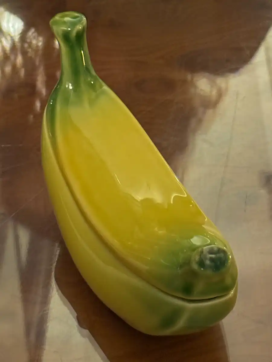 Ein keramischer Behälter, der wie eine gelbe Banane geformt und bemalt ist, steht auf einer polierten Holzoberfläche.