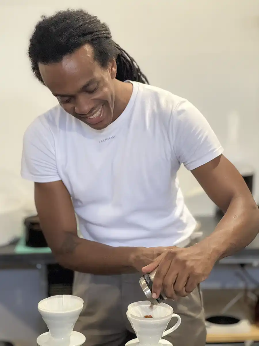 Geheimtipps-Martinique-Luigi-Germany Ein lächelnder Mann im weißen Hemd schüttet in einer hellen Küche Kaffeesatz in einen Pour-over-Filter.