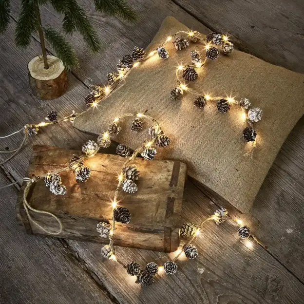 Lichterketten mit Tannenzapfen, arrangiert auf einem Kissen aus Sackleinen, einem Holztablett und einem rustikalen Holzboden.
