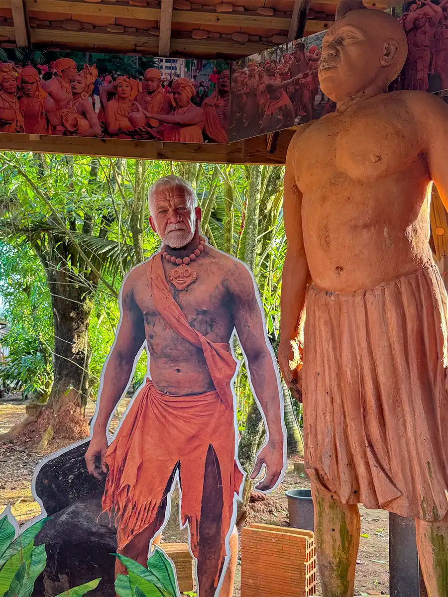 Martinique-Geheimtipps-Toepferdorf.-Homme-d-argiles-Figur Eine Pappfigur eines Mannes in Stammeskleidung steht neben einer Tonstatue unter einem Unterstand