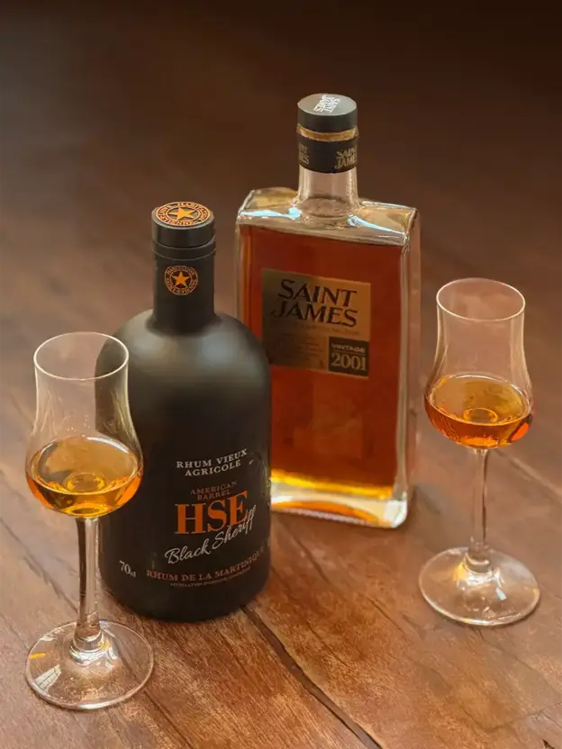Zwei Gläser Rum stehen neben Flaschen mit HSE Black Sheriff und Saint James Rum auf einem Holztisch.