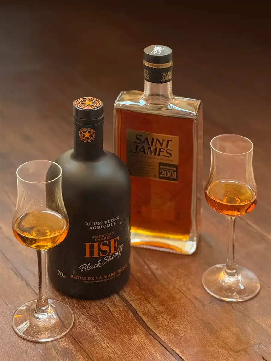 Zwei Gläser Rum stehen neben Flaschen mit HSE Black Sheriff und Saint James Rum auf einem Holztisch.