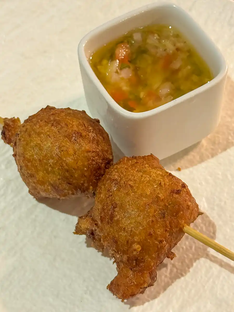 Martinique-Spezialitaeten-Accras Zwei goldbraun gebratene Bällchen auf einem Spieß mit einer kleinen Tasse Dip-Sauce auf einer weißen Unterlage.