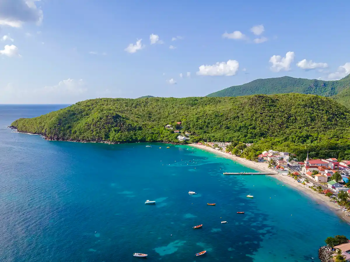 Martinique-Straende-Anse-d-Arlet Luftaufnahme einer üppig grünen Küstenlinie, eines Sandstrandes und von Booten, die auf dem klaren türkisfarbenen Wasser treiben.