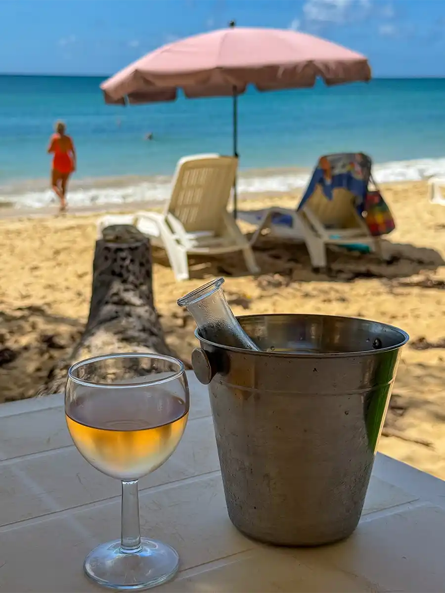 Martinique-Straende-Les-Salines-Rose Ein Glas Weißwein und ein Weinkübel auf einem Tisch, mit einem Strand und einem Sonnenschirm im Hintergrund.