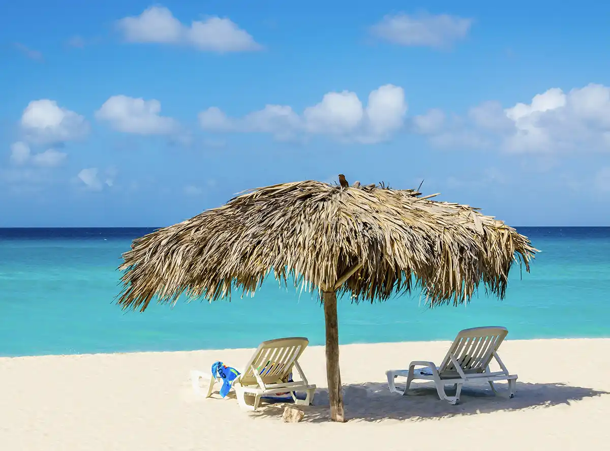 Martinique-Strand-Karibik Zwei Liegestühle unter einem strohgedeckten Sonnenschirm an einem Sandstrand mit kristallklarem, blauem Meerwasser unter einem sonnigen Himmel.