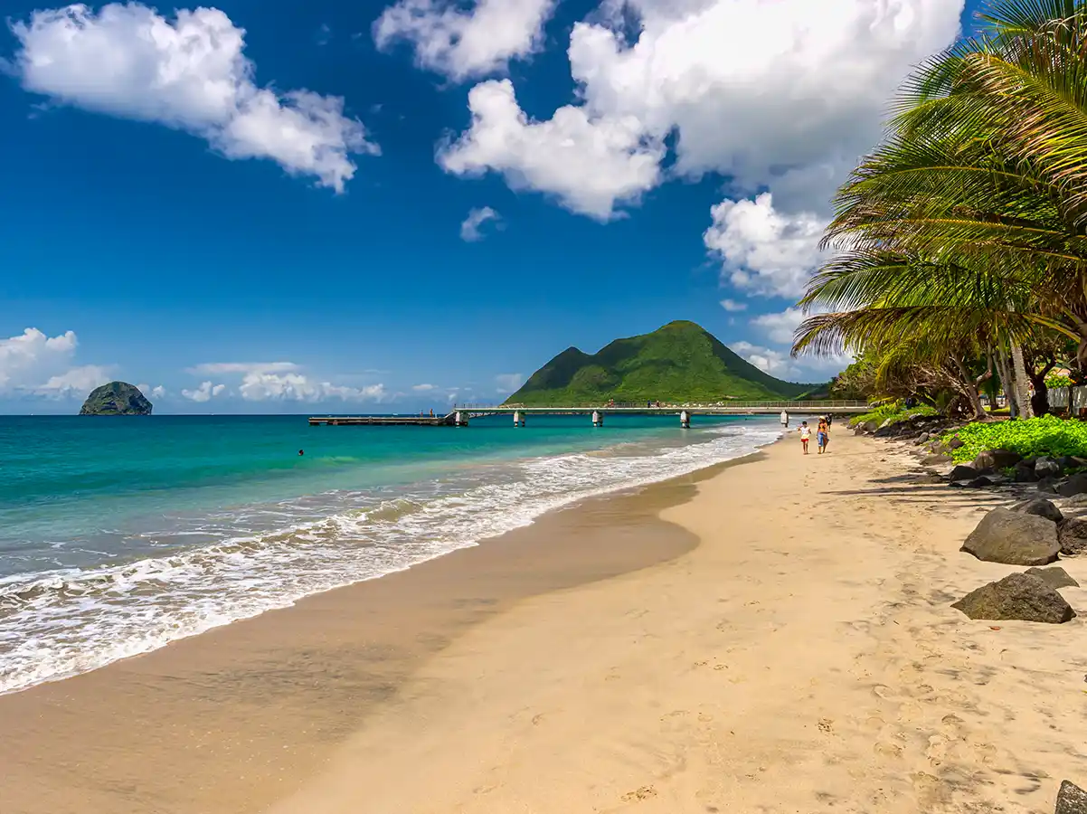 Martinique-Strand-Le-Diamant Sandstrand mit Palmen, türkisfarbenes Wasser, ein Pier und grüne Berge unter einem blauen Himmel mit Wolken.