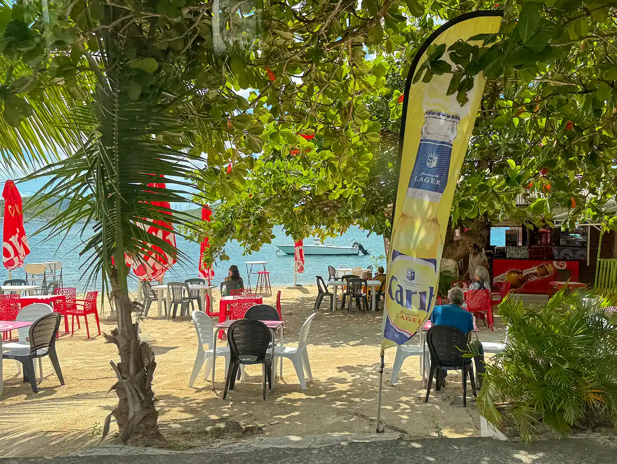 Martinique-Strand-Le-Marin Strandcafé im Freien mit rot-weißen Stühlen, Sonnenschirmen und einer Carib Lager-Flagge in Meeresnähe unter Laubbäumen.
