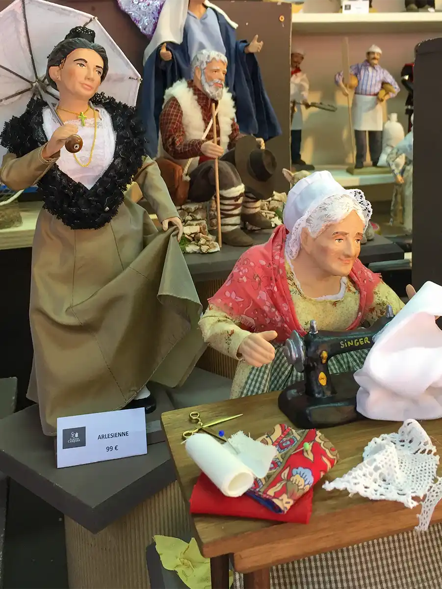 Santons-Arlesienne-Weihnachtsmarkt Zwei traditionelle Puppen: eine steht mit einem Regenschirm, die andere näht an einer Maschine mit Stoffen auf dem Tisch.