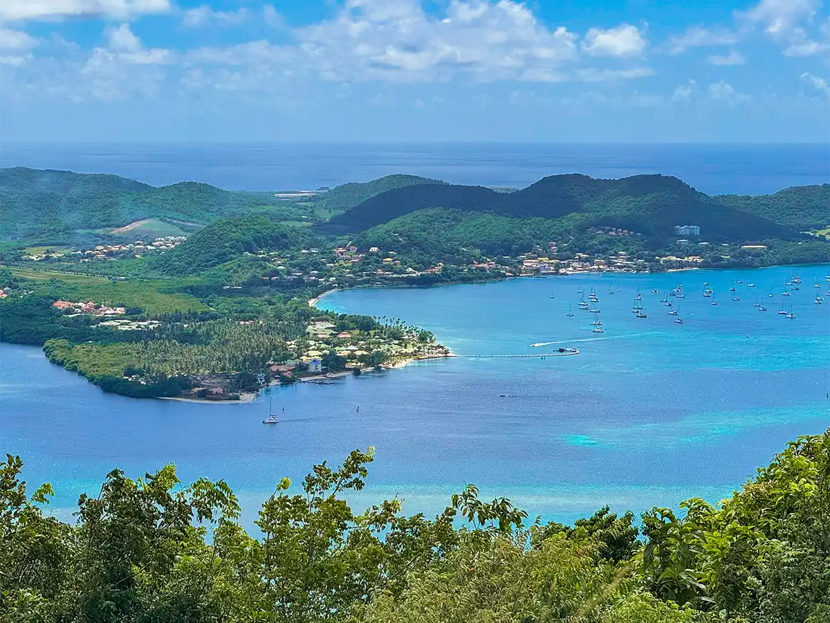 Schoenste-Straende-Martinique-Saint-Anne Luftaufnahme einer Bucht mit türkisfarbenem Wasser, Segelbooten und üppig grünen Hügeln unter einem blauen Himmel mit vereinzelten Wolken.